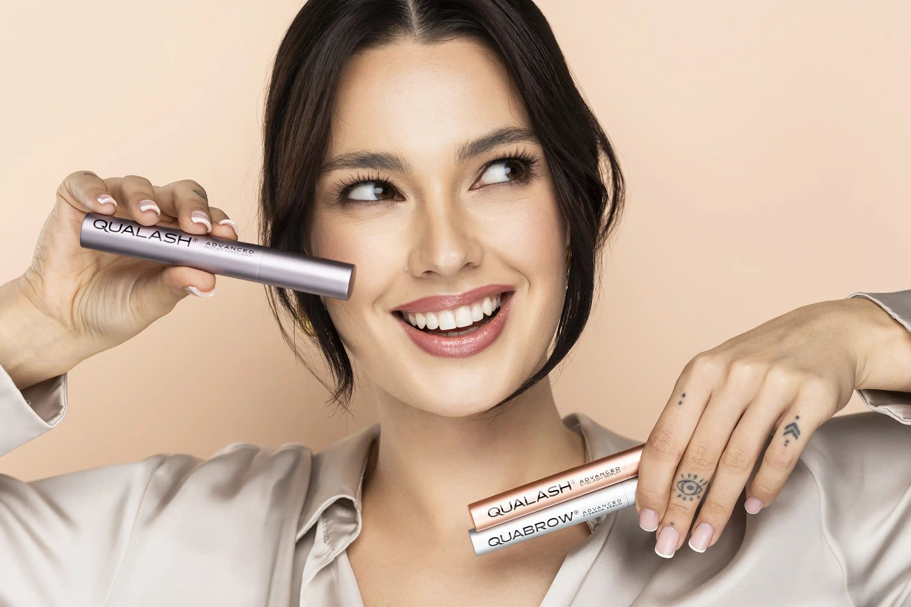Smiling woman holding QUALASH Mascara and QUABROW & QUALASH Serums – natural makeup look