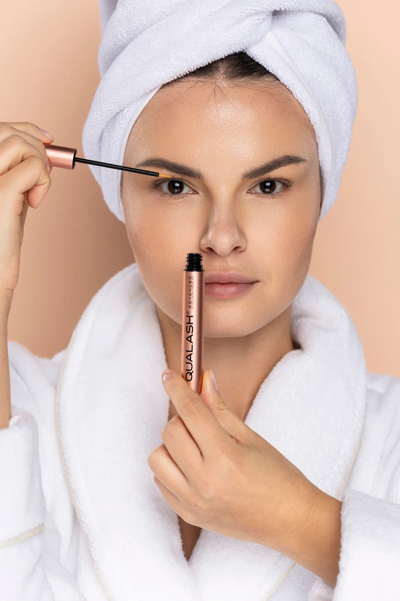 Front-facing beauty portrait of a woman holding a Qualash cosmetic tube and applying brow serum with an applicator on a neutral background