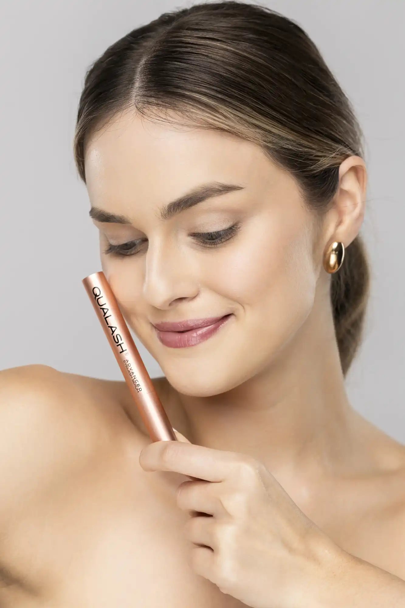 A woman with long brown hair wearing a gold earring, holding a rose gold tube of mascara.