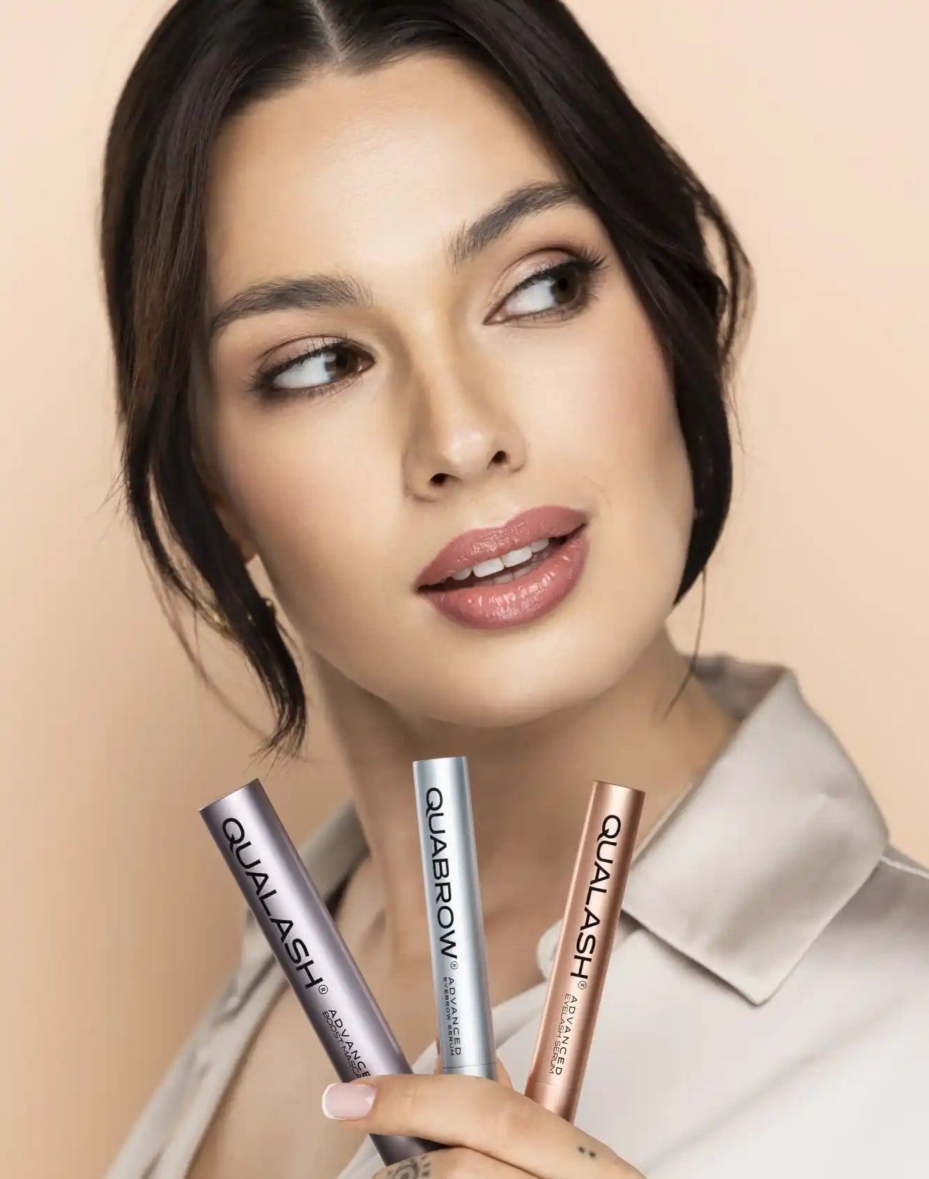 Beauty portrait of a woman holding three cosmetic tubes labeled Qualash and Quabrow against a soft neutral background